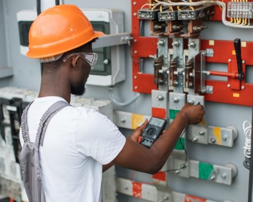 African american electrician in safety helmet and glasses checking voltage in switchgear while working at solar station. Repairing of modern equipment. Production of green energy.